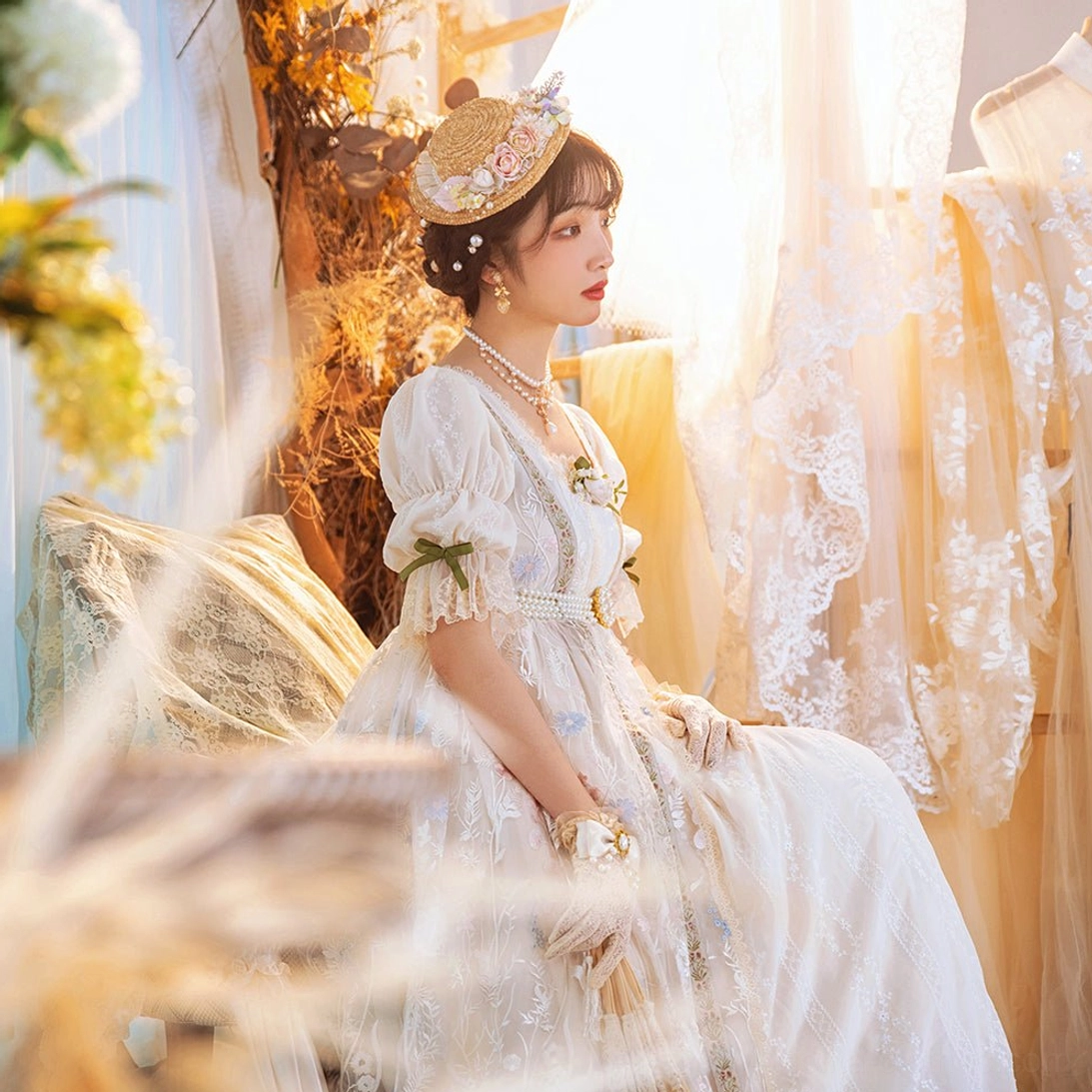 Seated vintage lolita full-body portrait in ivory ruffle dress straw bonnet pearl necklace and retro elegant makeup indoor boudoir style room setting