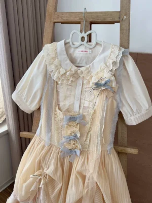 Sweet Lolita blouse coordinate with lace Peter Pan collar and beige jumperskirt featuring blue lace bows, puff short sleeves, and ruffled front placket design on wooden hanger display