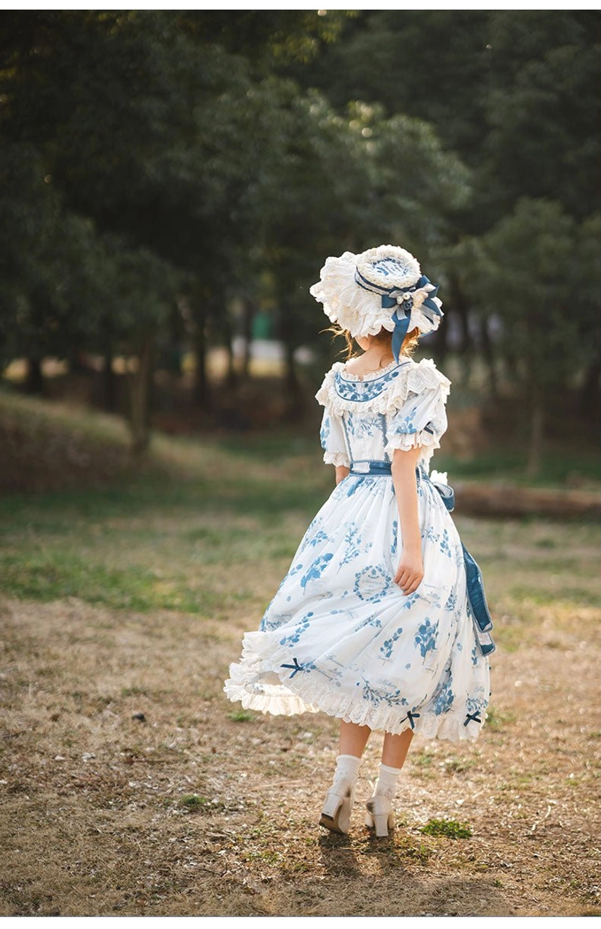 Rear full-body view of short-sleeve blue and white printed Lolita one-piece dress with ruffled round neckline, tiered midi skirt, and matching straw bonnet with blue trim, photographed on woodland dirt ground