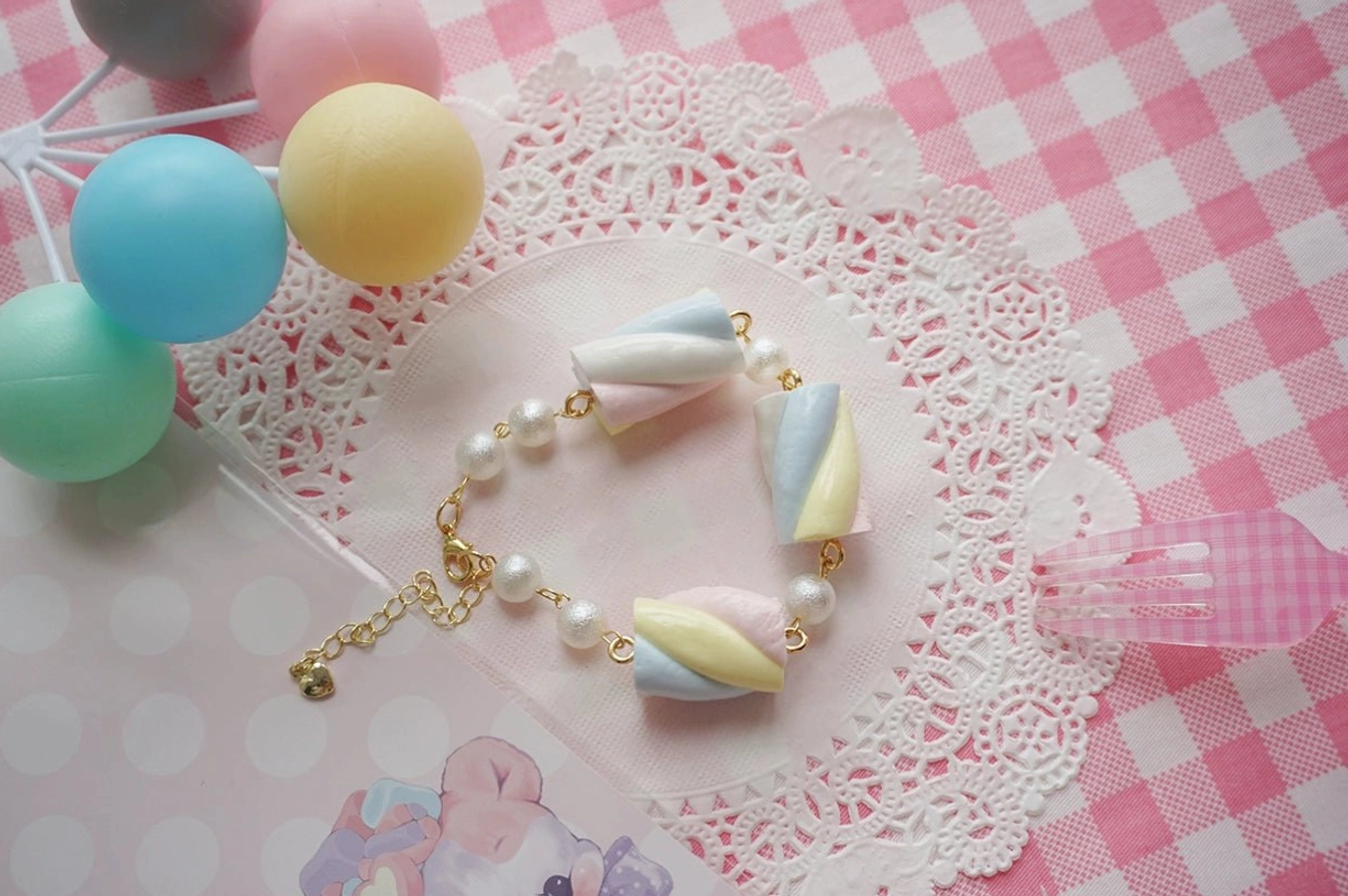 Single pastel cotton candy resin lolita bracelet on checkered tablecloth with lace doily background, featuring compact marshmallow string design with gold extension chain and cute pastel color scheme