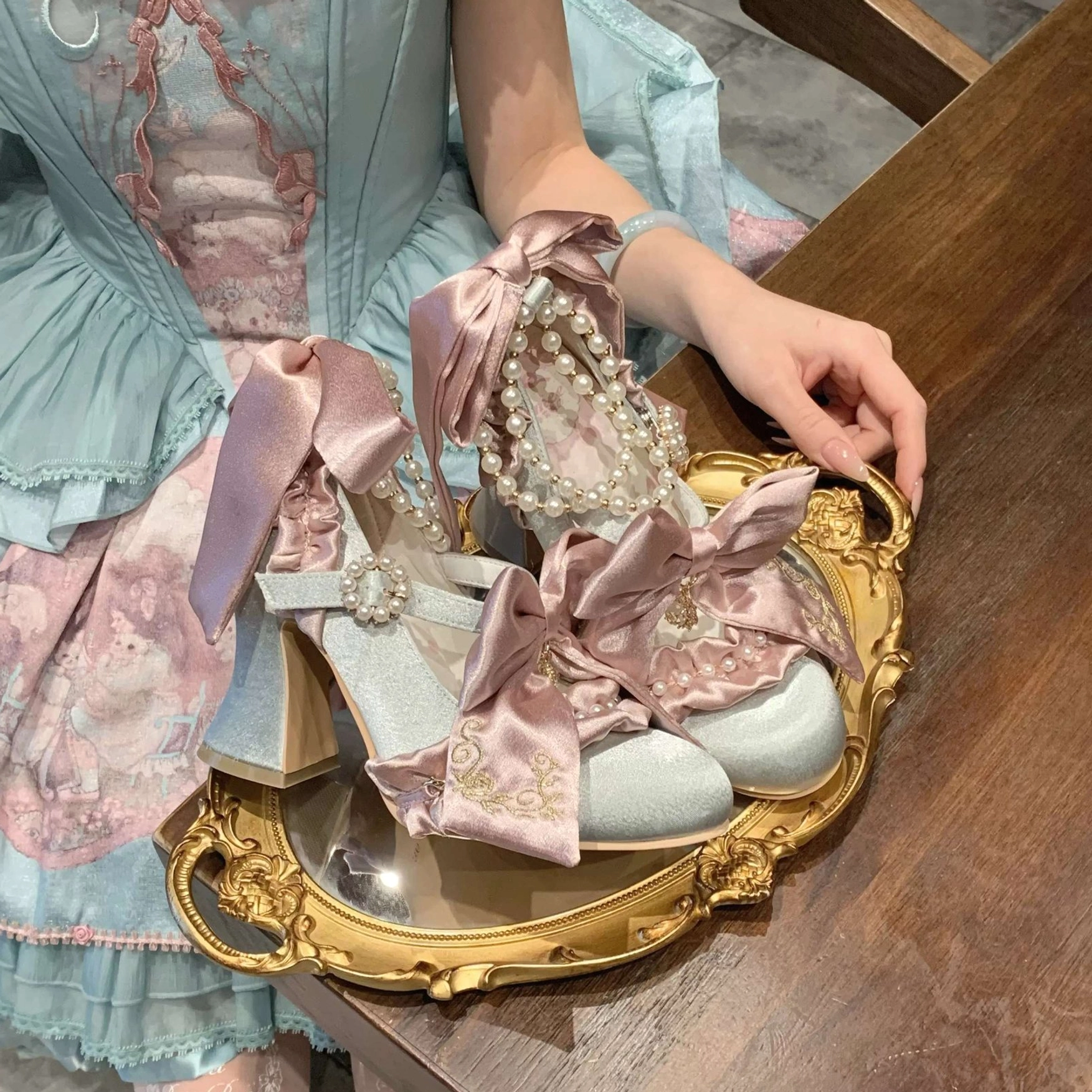 Sky-blue Classic Lolita block heel shoes with pink satin bows and pearl decorations, flat lay on golden decorative tray with blue-pink print Lolita dress styling and shiny satin fabric surface