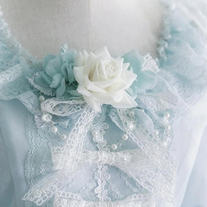 Close-up flat lay view of sky blue Lolita collar accessory with white artificial rose, ruffled lace trim, and dangling pearl chain details placed on light blue sheer fabric background