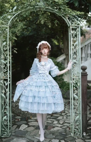 Full-body frontal view of sky blue tiered ruffle Lolita OP dress with matching pearl-adorned sweet lolita BNT headwear, displayed on model at garden iron archway setting with off-the-shoulder design and white stockings