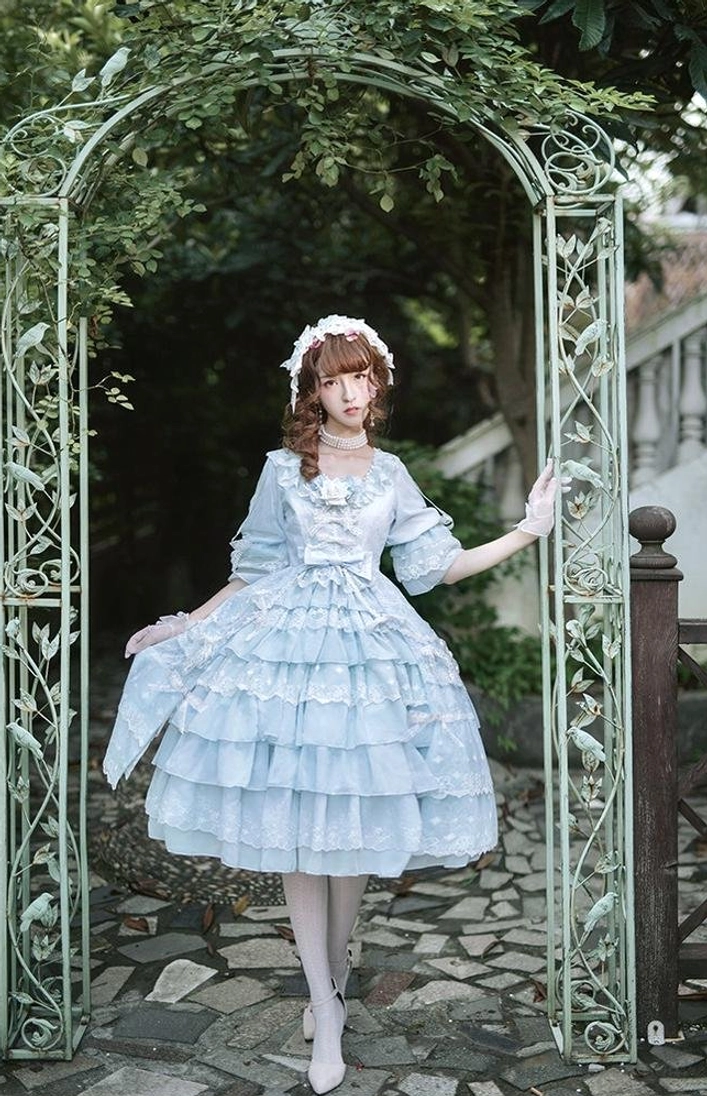 Full-body frontal view of sky blue tiered ruffle Lolita OP dress with matching pearl-adorned sweet lolita BNT headwear, displayed on model at garden iron archway setting with off-the-shoulder design and white stockings
