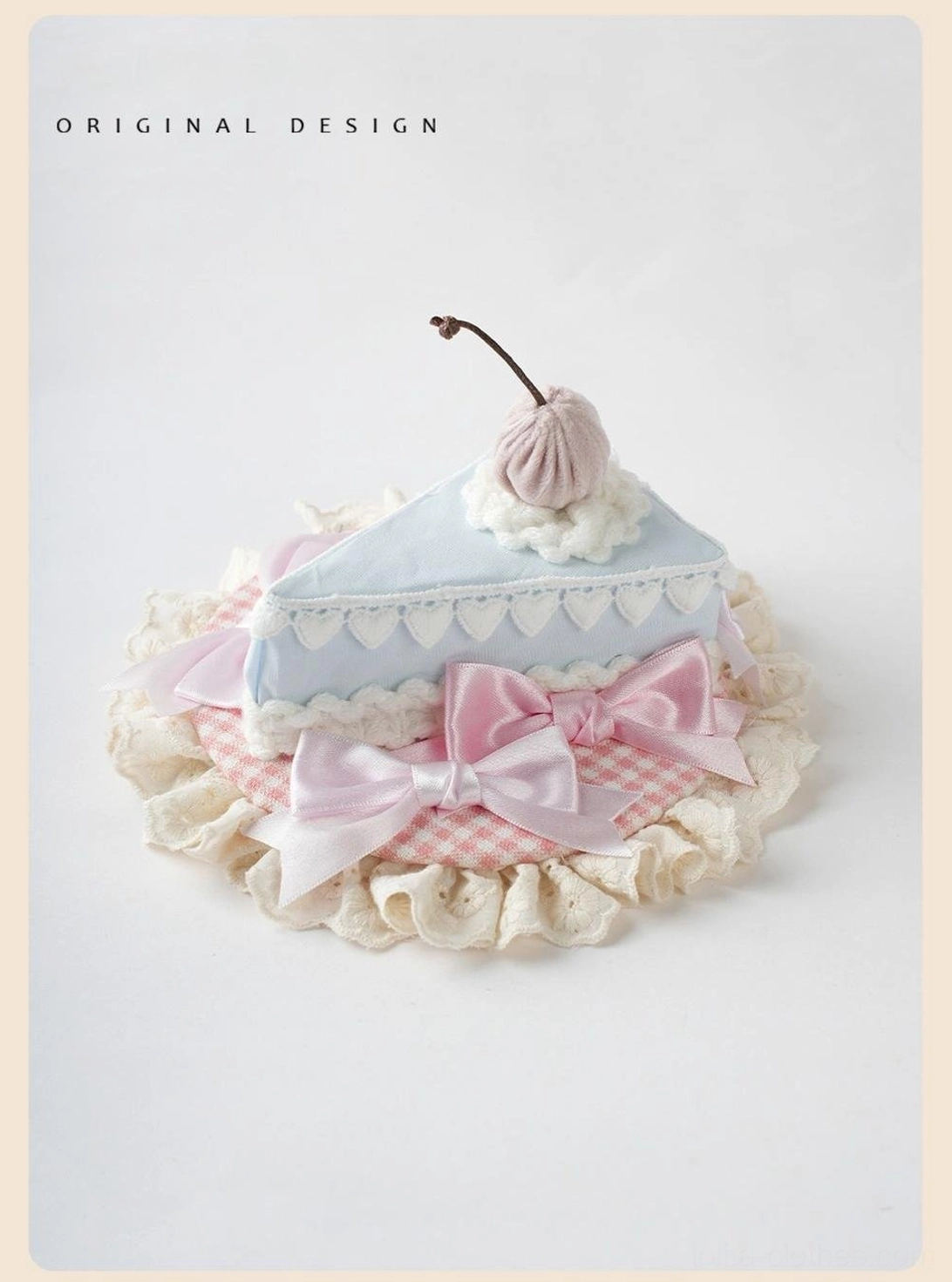 Slice cake style sweet lolita hat fascinator with light blue cake slice base, pink gingham underlayer, pink satin bows, ruffled lace trim, cherry cream topper, flat lay