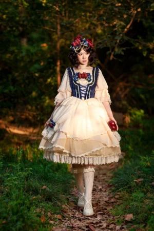 Snow White split-style Lolita OP dress with navy corset and cream tiered skirt, front view on model in outdoor forest setting wearing matching hair accessory and holding red apple prop