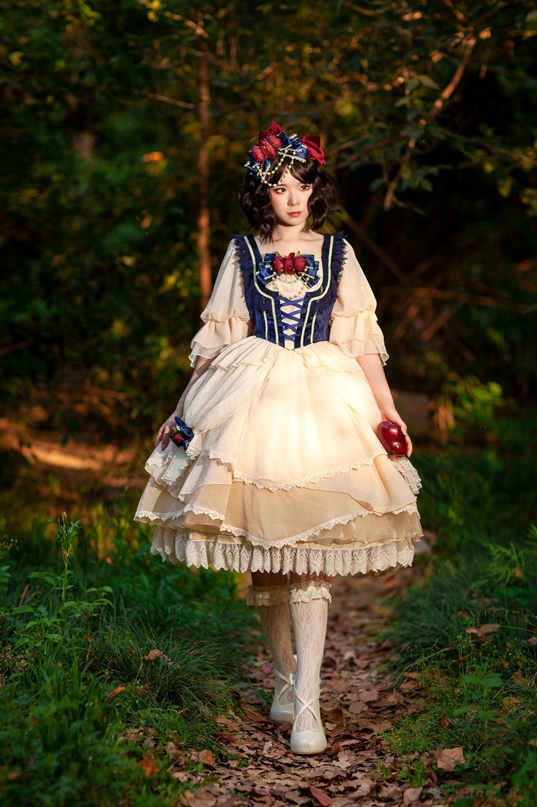 Snow White split-style Lolita OP dress with navy corset and cream tiered skirt, front view on model in outdoor forest setting wearing matching hair accessory and holding red apple prop
