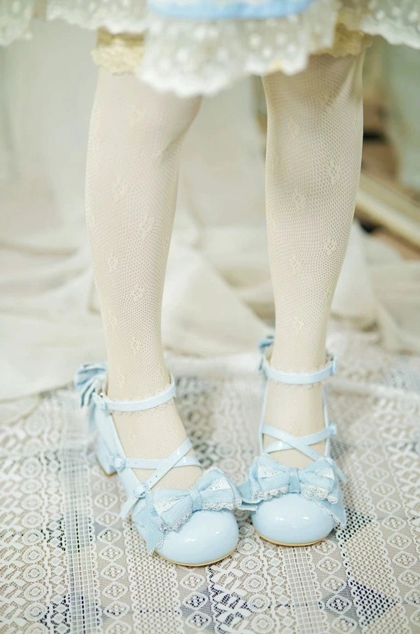 Soft blue patent sweet Lolita wedding heels worn on model's feet with ruffled hem skirt and patterned stockings on lace floor mat, delicate floral decorations and cross ankle straps visible