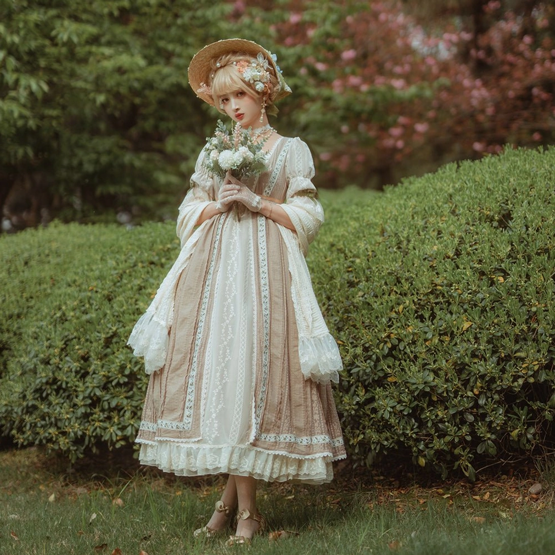 Standing vintage lolita full-body shot wearing tan ruffled dress matching shawl straw bonnet and small flower bouquet in outdoor garden hedge background