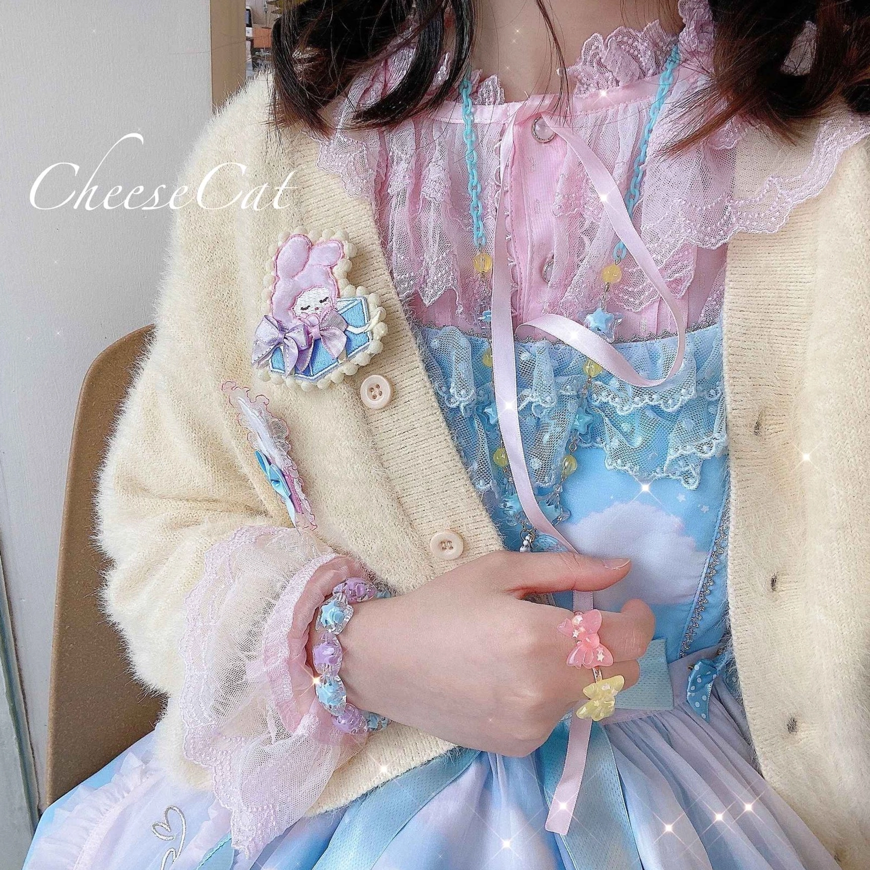 Close-up upper-body shot of girl in sky-blue pink Lolita dress with cream knit cardigan wearing star beaded Lolita bracelet and matching rings with cute cartoon brooch, showing daily Lolita coordinate