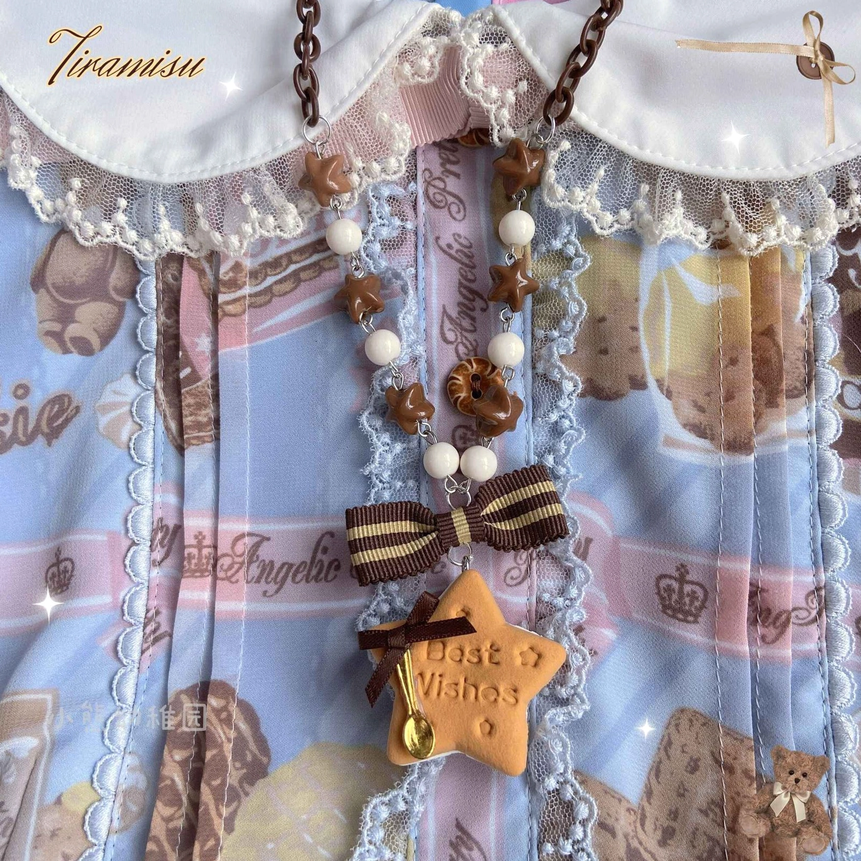 Close-up of star-shaped cookie lolita necklace worn on blue lace-trimmed dress collar with crown and dessert prints, featuring striped brown bow, star biscuit charm with Best Wishes text, and mixed beaded chain