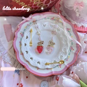 Two strawberry charlotte lolita necklaces styled together on stacked ceramic plates with gingham fabric and pink ribbon props showcasing beaded chain construction and gold hardware