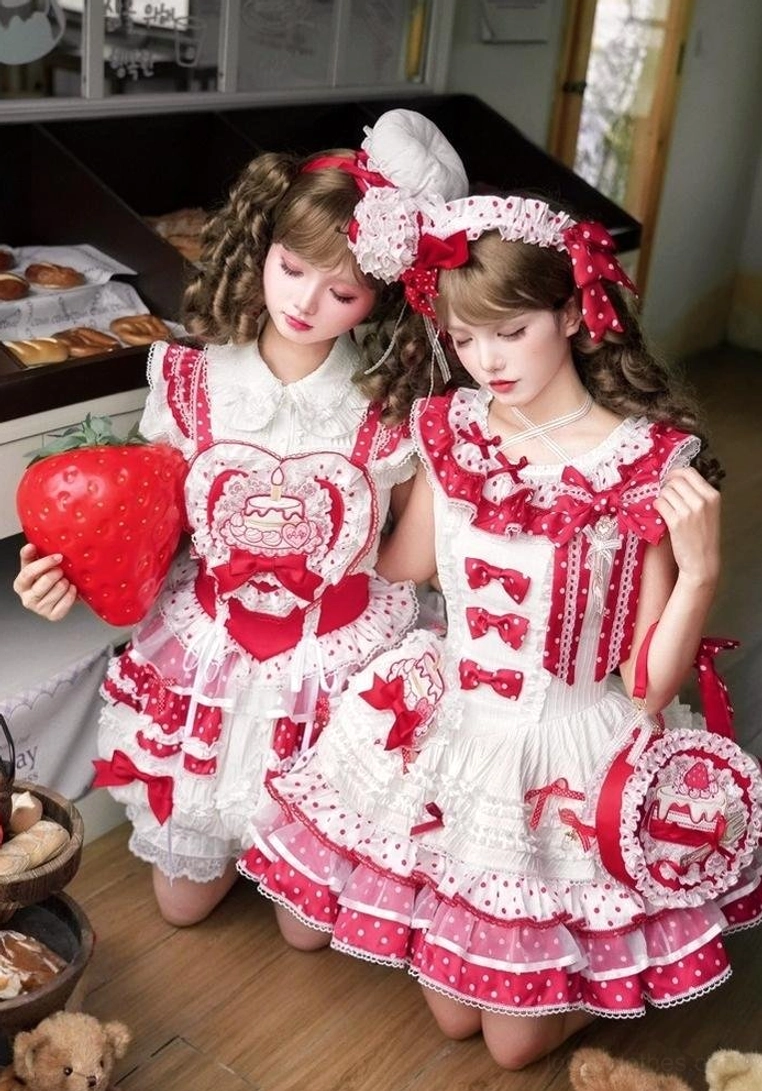 Two lolita models in kneeling half-body pose with strawberry-themed sweet lolita dress featuring heart bear embroidery, red polka dot layers, and large strawberry prop