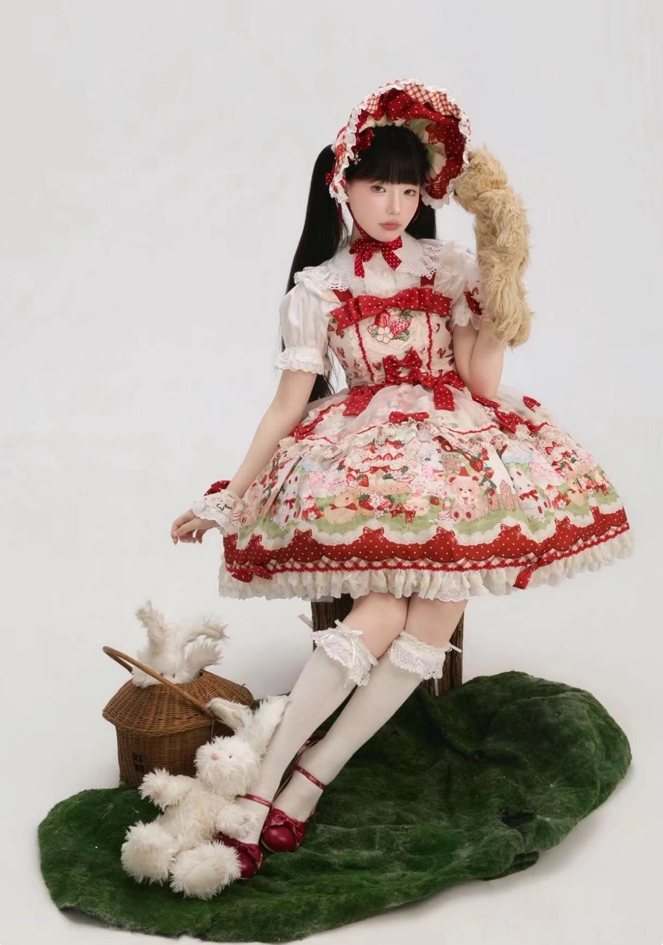 Strawberry Lolita JSK dress with big hair bow sitting side shot on woodland decor green mat, wearing white lace stockings with red straps, holding plush prop, demonstrating comfortable seated fit and A-line silhouette