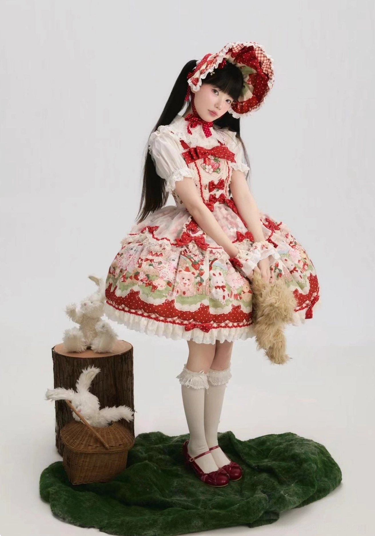 Strawberry themed sweet Lolita JSK dress three-quarter front side angle with matching bonnet, held with plush bear prop, scalloped lace hem, decorative neck bow, red leather Mary Jane shoes, woodland decor setting