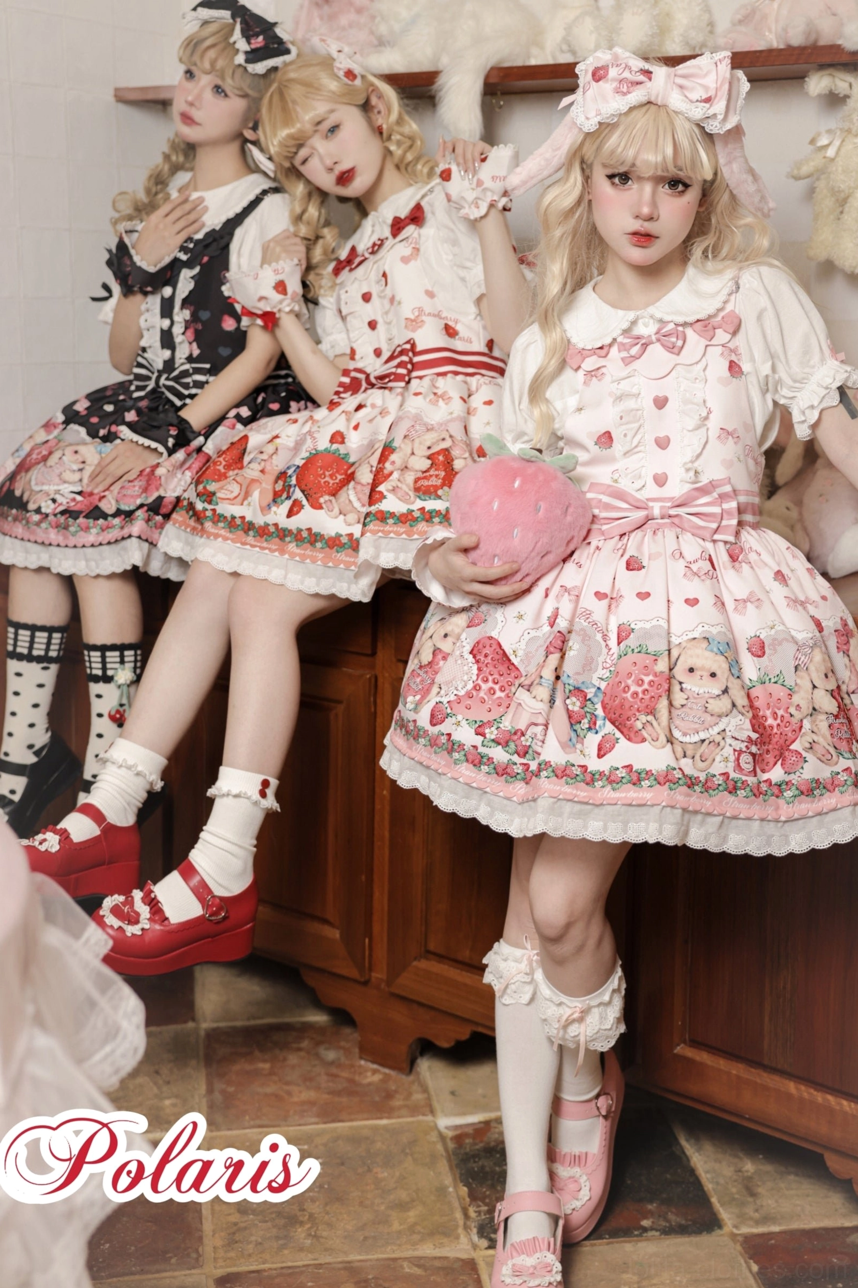 Three sweet lolita models wearing different colorways of strawberry print salopette dresses with coordinating hair bows and mary jane shoes in kitchen