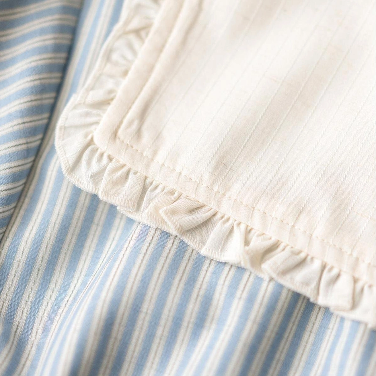 Fabric texture close-up of blue and white vertical striped woven material with ruffled white cotton edge trim, highlighting soft delicate textile details