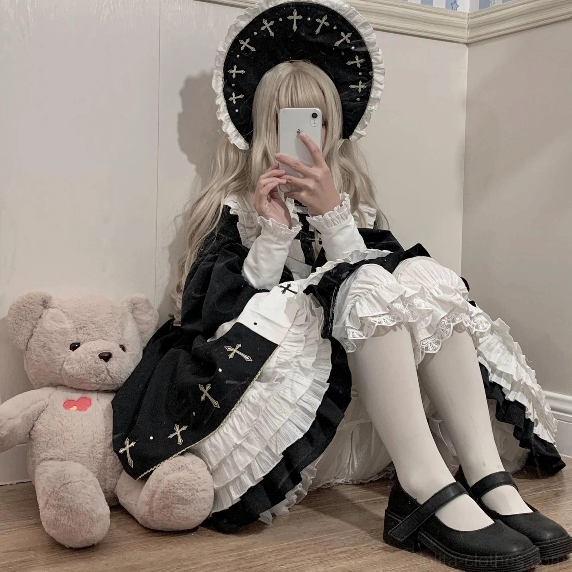 Seated lolita model taking mirror selfie wearing cross-decorated nun bonnet cross-print op dress white stockings black mary jane flats white lace bloomers partially visible