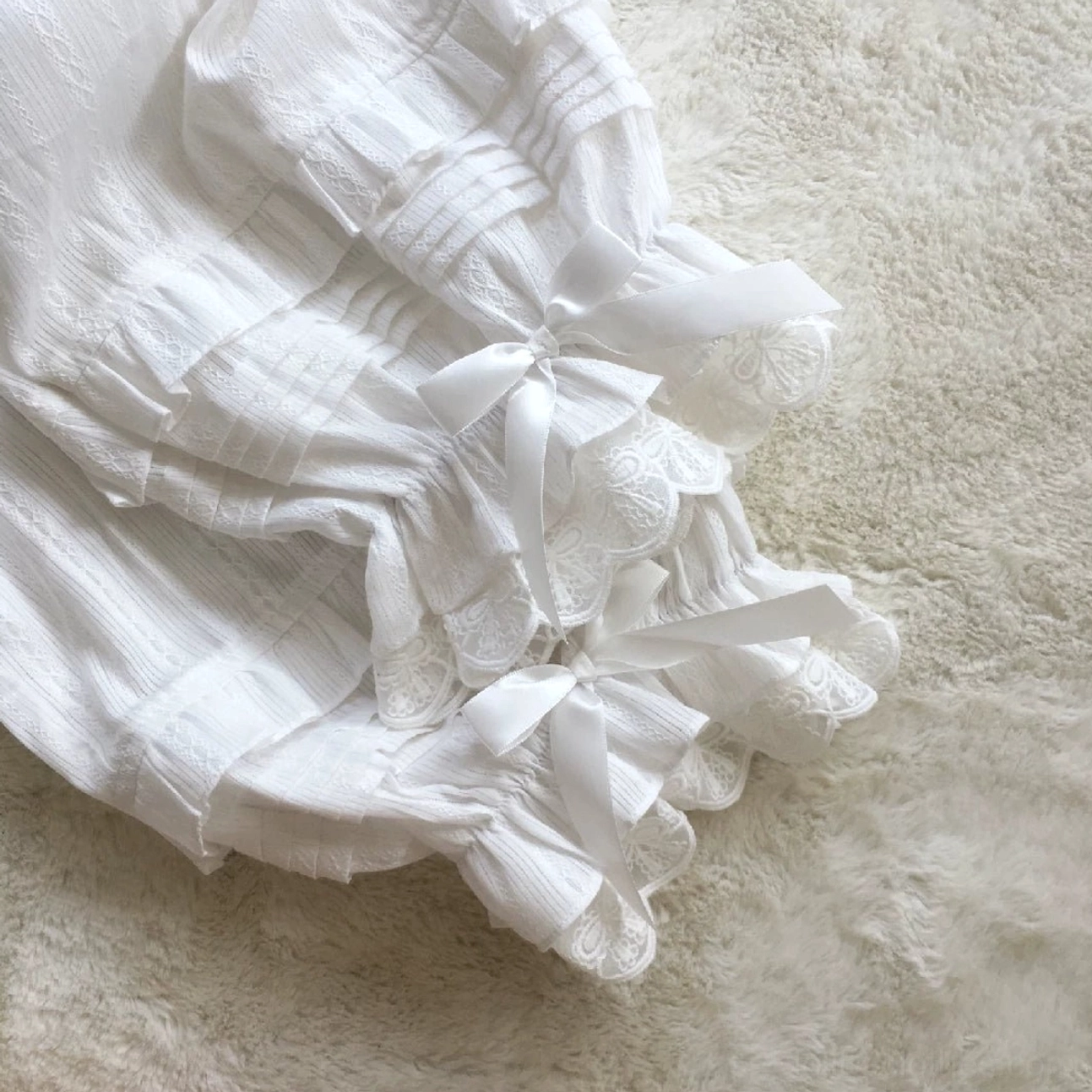 Close-up angled flat lay detail view of white sweet lolita bloomers cuffs featuring multiple satin ribbon bows lace ruffled hem accordion pleated fabric jacquard texture