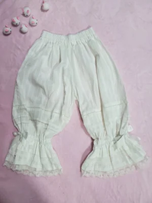 Sweet Lolita cotton bloomers with vertical stripes, white lace ruffled cuffs, and elastic waistband displayed on pink fabric background with decorative dolls—flat lay overview of classic Lolita undergarment