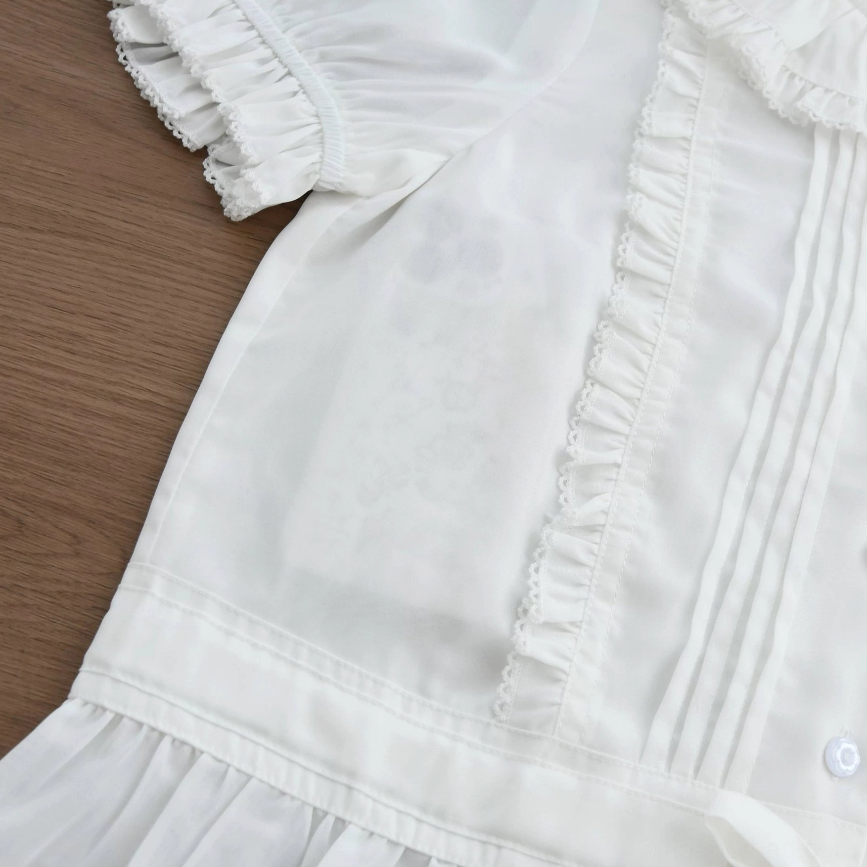 Flat lay detail view of sweet Lolita blouse on wooden surface showing white fabric, front pintuck panels with lace frills, ruffled sleeve edge, layered ruffle hem details