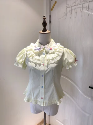 Sweet lolita short-sleeve blouse with pale yellow coloring, lace-trimmed ruffled Peter Pan collar, puffed short sleeves, front colorful candy buttons and hem ruffle trim displayed on mannequin with wooden door backdrop