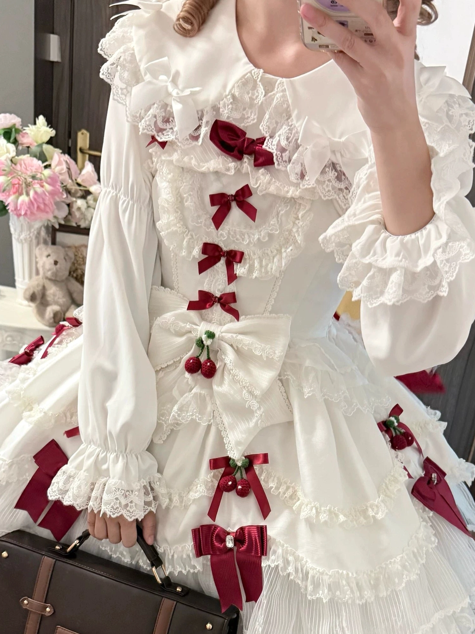 Side mirror selfie of model in sweet Lolita dress featuring red bows and cherry-shaped ornaments on large front bow with white Peter Pan collar blouse