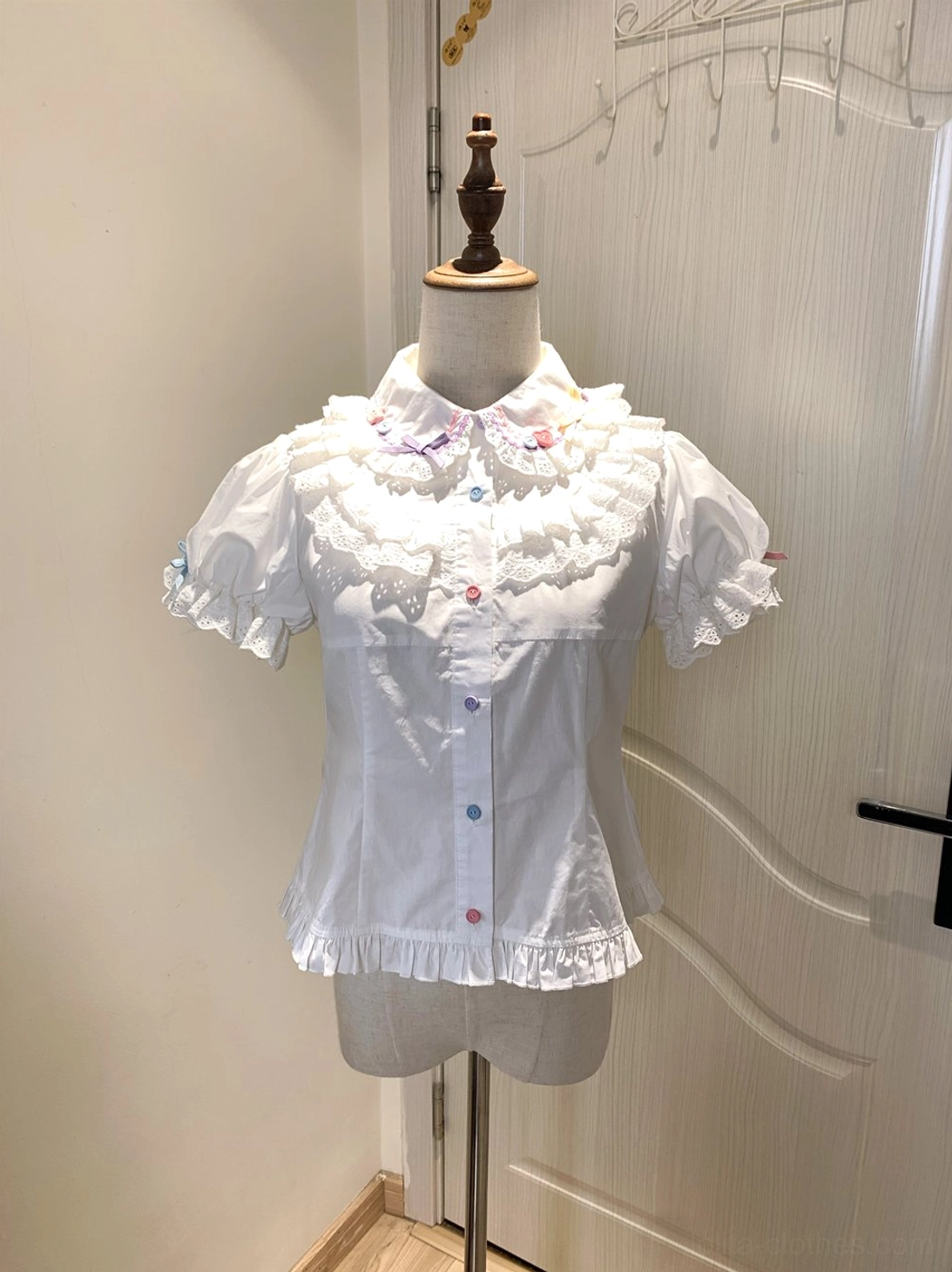 Pure white sweet lolita short-sleeve blouse with lace ruffled Peter Pan collar puffed sleeves colorful buttons and hem ruffle trim displayed on mannequin studio backdrop