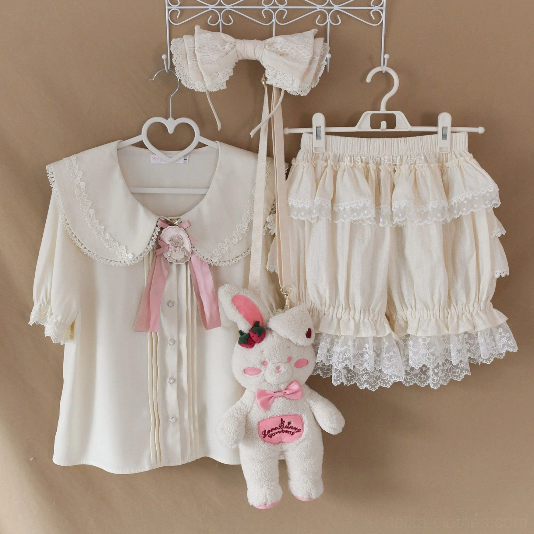 Sweet lolita coordinate set with white blouse featuring doll collar and pink bow brooch, bloomer shorts with ruffle and lace trim, hanging flat lay retail display with plush rabbit prop