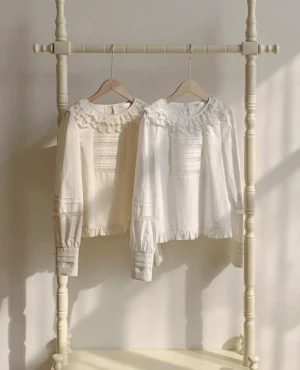 Sweet lolita cotton blouse with ruffled lace round collar and front pintuck details displayed on wooden clothing rack with warm indoor lighting