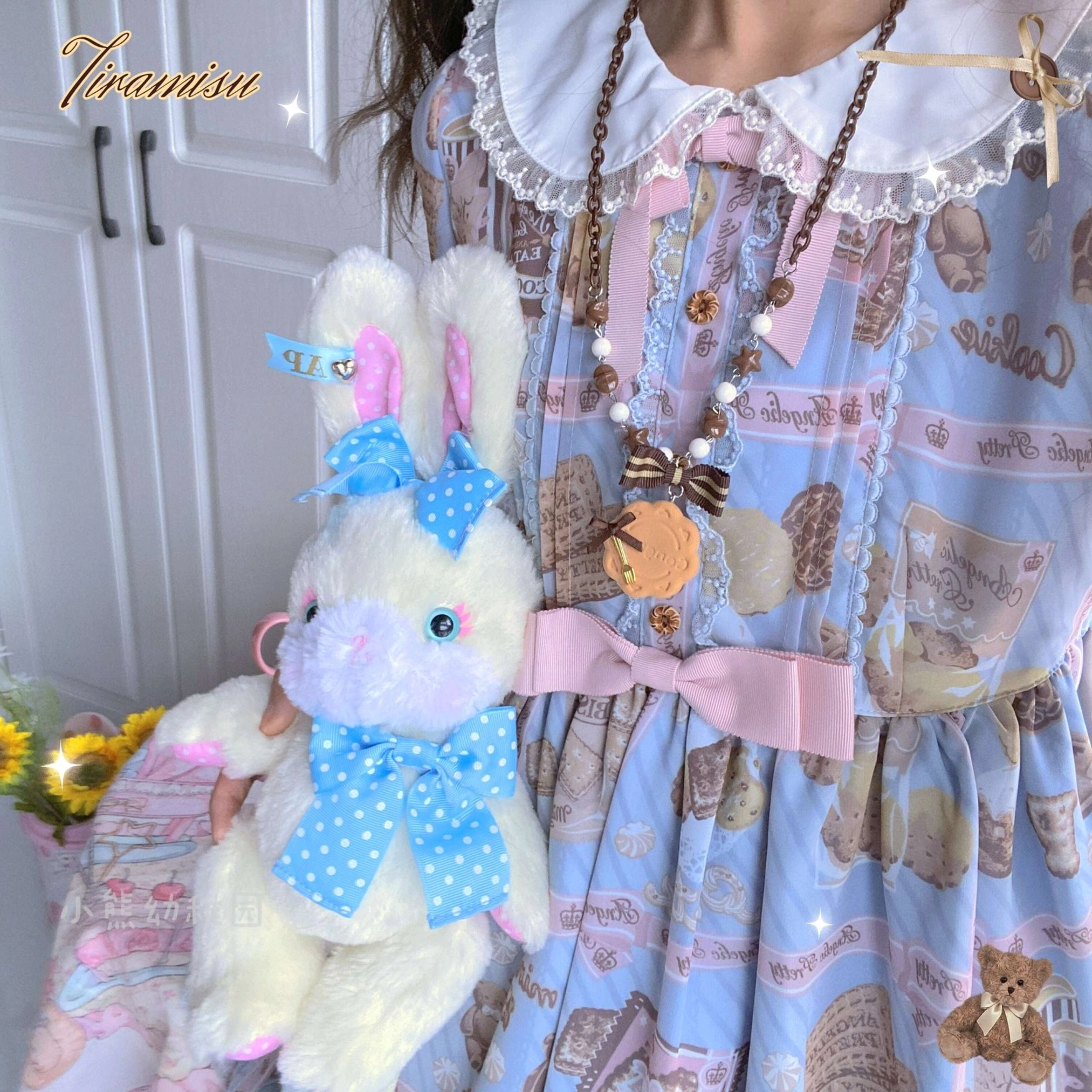 Styled outfit photo of girl wearing blue lolita dress with dessert and teddy bear prints paired with matching biscuit charm necklace, coordinated with blue bowknot plush rabbit doll on white door background