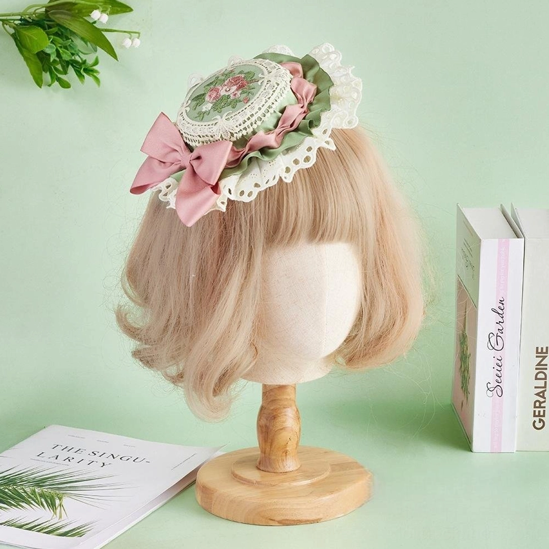 Sweet Lolita floral embroidery top hat with pink satin bow and white ruffled lace trim, worn on blonde mannequin displaying ornate round floral medallion with sage green accents