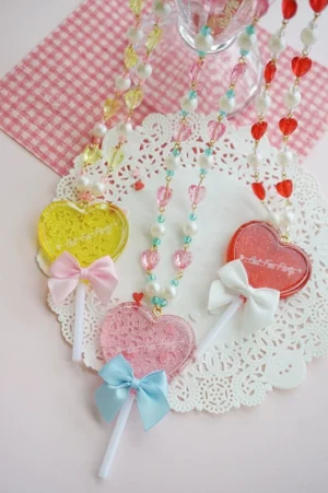 Overhead flat lay view of multiple sweet lolita heart-shaped lollipop necklaces in various pastel colors displayed on gingham placemat with white lace doily and glass prop