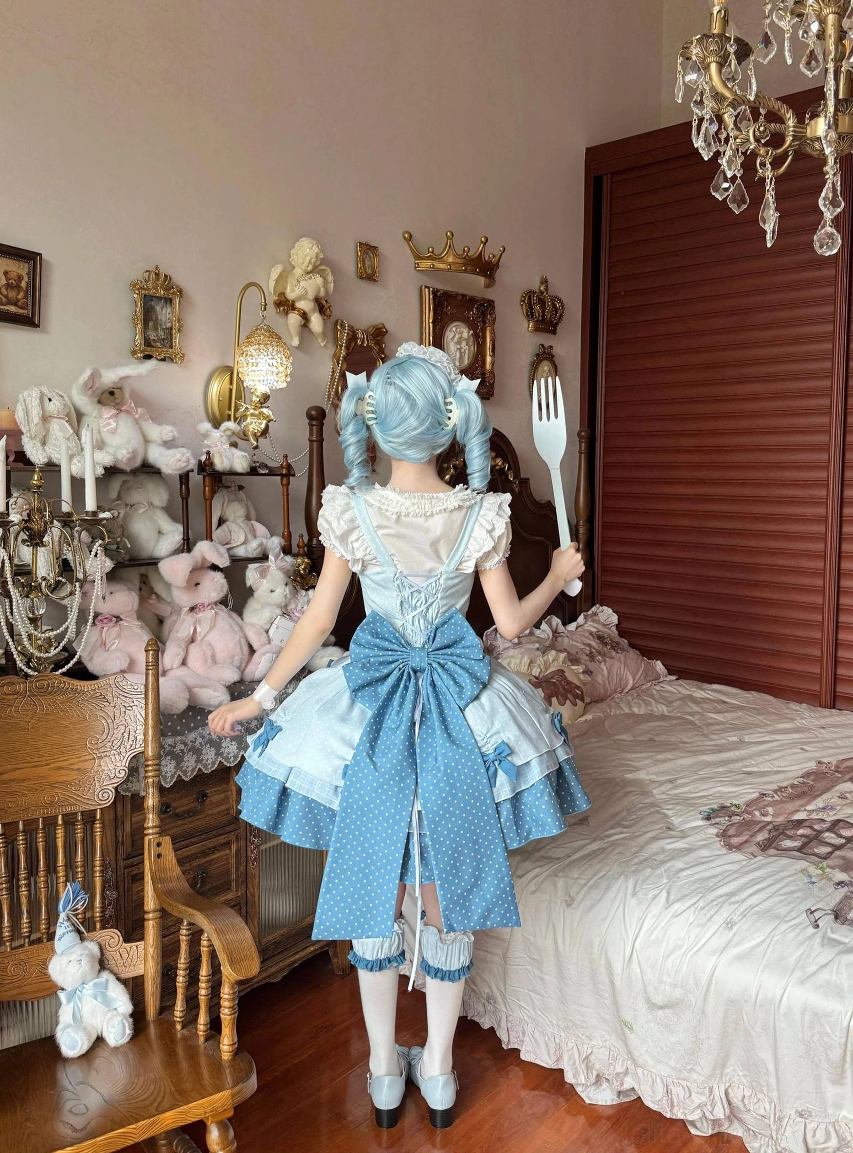 Back view of Sweet Lolita dress worn by model showing large blue back waist bow, polka dot skirt trim details, tiered ruffled construction, model holding fork prop, vintage bedroom setting