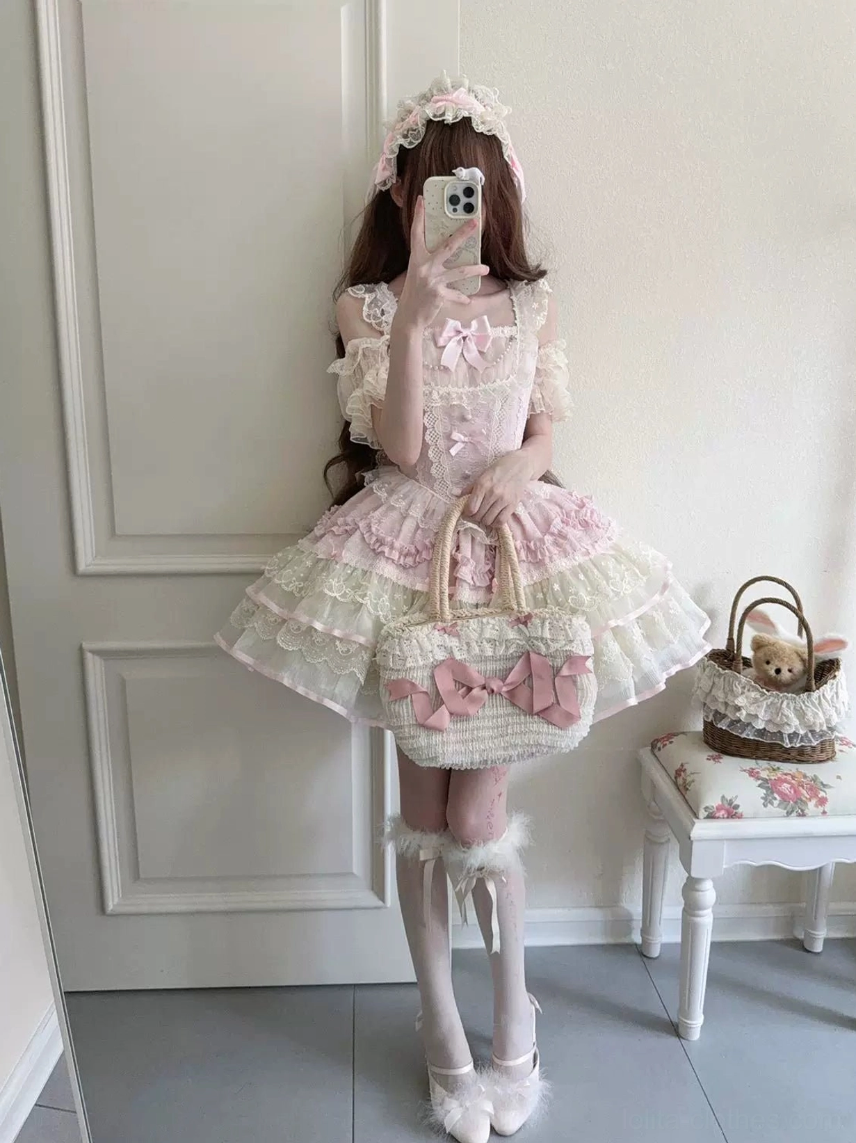 Front mirror selfie featuring female model in pink and ivory sweet lolita jumper skirt with hair flower accessory, fitted bodice, tiered lace petal ruffles, fluffy leg warmers, ballet flats, and matching lolita handbag