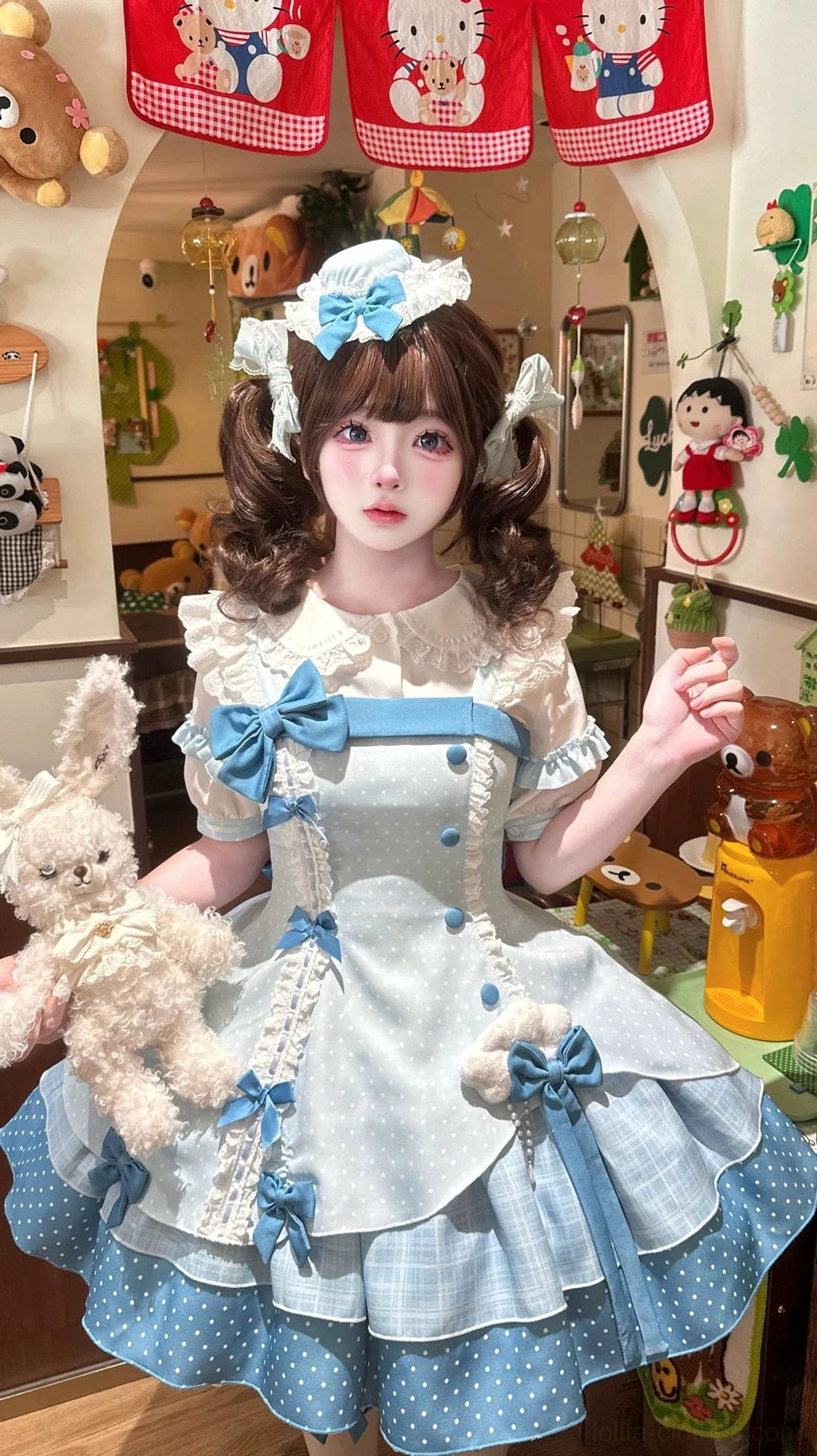 Sweet Lolita JSK dress worn by model front three-quarter angle view in doll collection bedroom, wearing matching hat, leg garters, oversized blue bows, carrying plush rabbit accessory