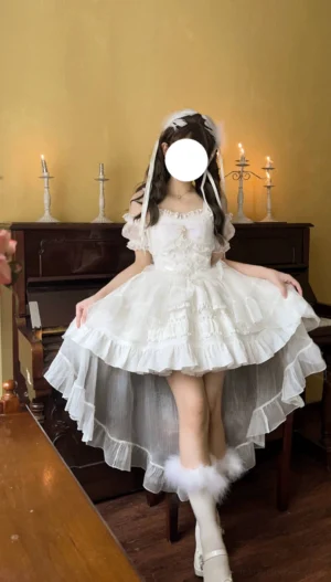 White high-low hem sweet Lolita JSK dress front three-quarter view with off-shoulder short sleeves, ruffled tulle layered skirt, lace-trimmed bodice, detachable bow, and coordinating white leg warmers in vintage piano room setting