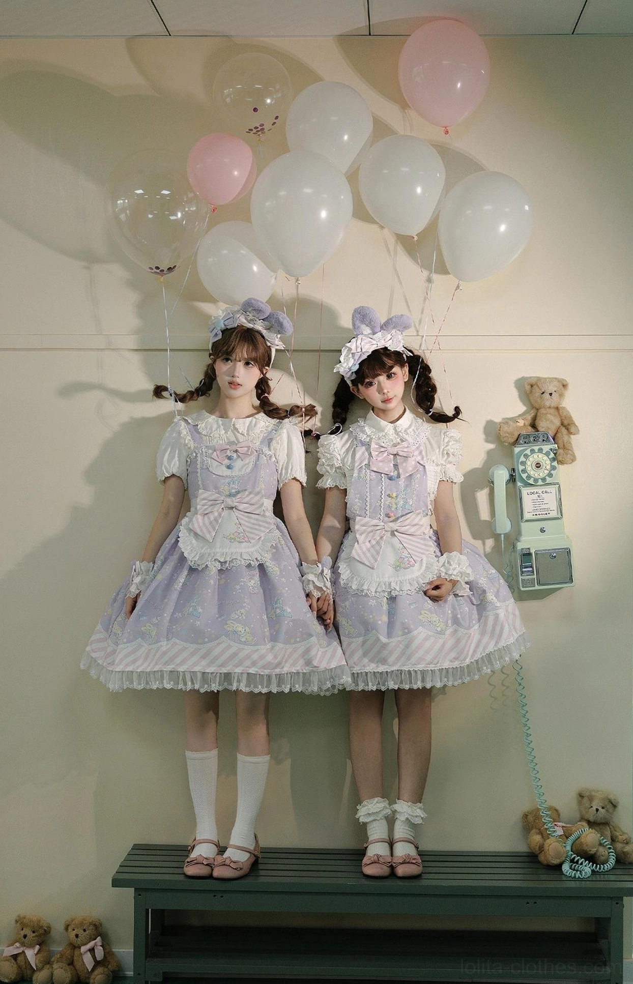 Full-body front standing view of pastel purple and light pink sweet Lolita dresses with matching bunny ear headwear on models sitting on bench, showing ruffled lace apron front, delicate bow details, balloon backdrop with plush toys
