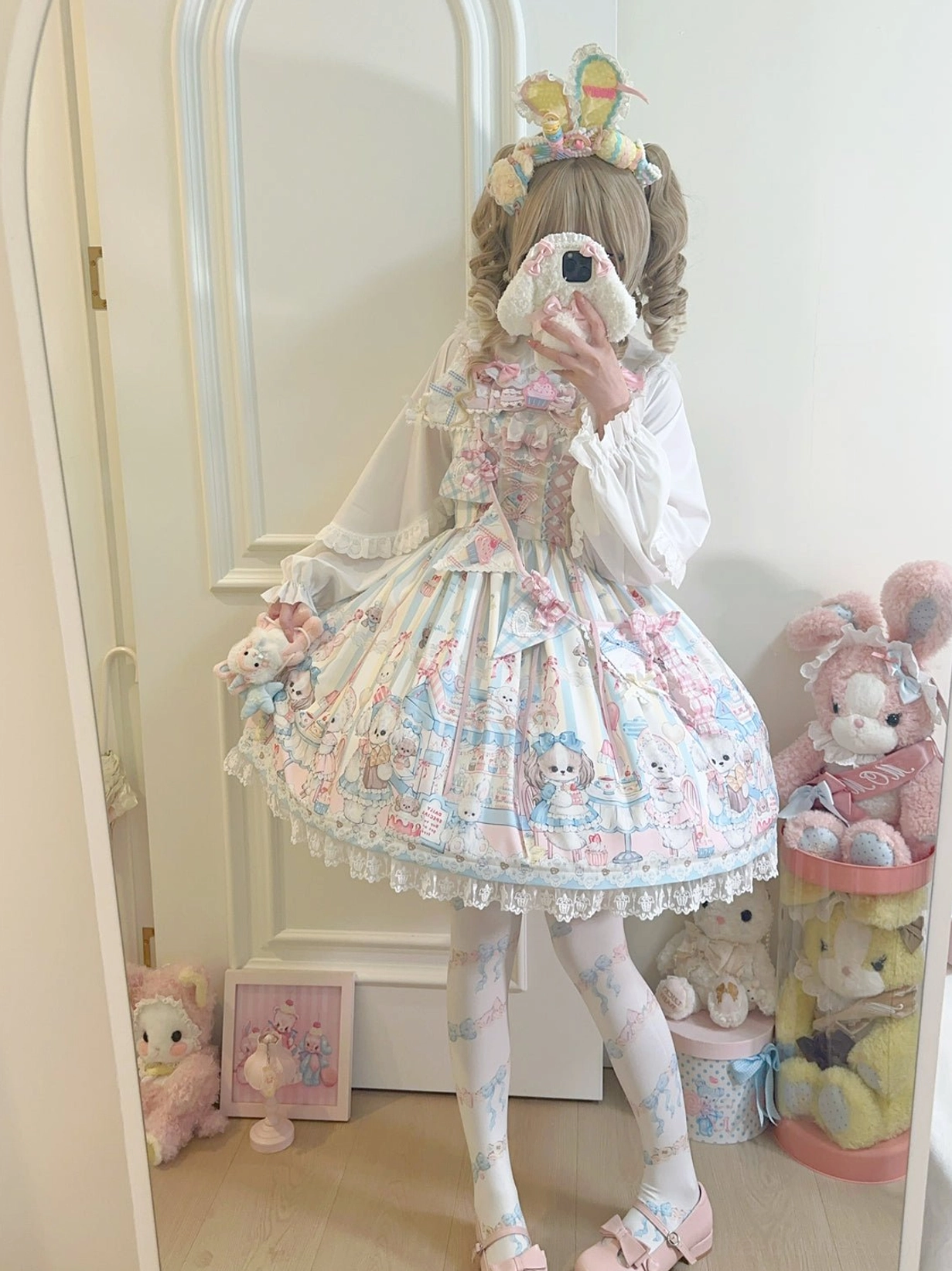 Sweet Lolita girl wearing puppy print JSK jumper skirt three-quarter front mirror view posing with hand near chin, cozy indoor setting with plush toys and framed wall decor complete kawaii aesthetic