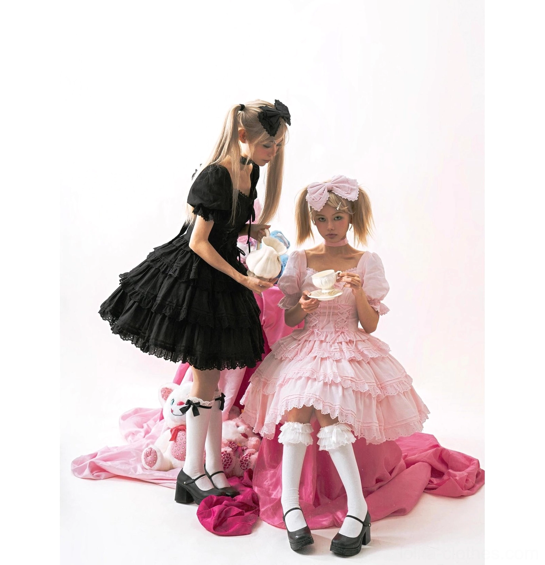 Black and pink sweet lolita one-piece dresses with tiered ruffled skirts, matching hair bows, short puff sleeves, worn by two models in tea party themed pose with formal lolita footwear
