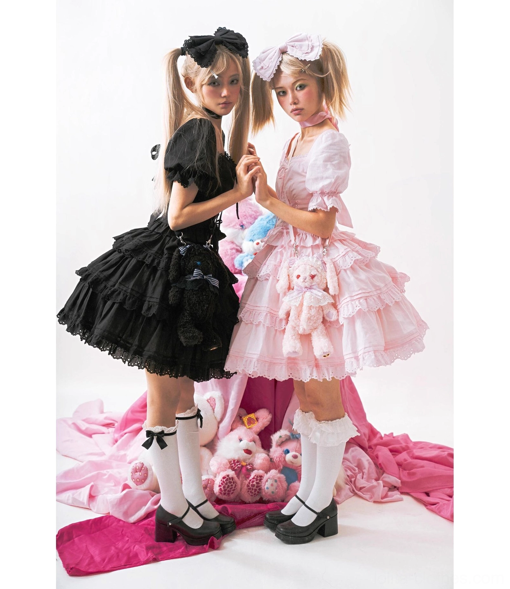 Matching sweet lolita one-piece dresses in black and pink with tiered ruffled skirts, puff sleeves, and coordinating hair bows worn by two models with studio props and Mary Jane shoes
