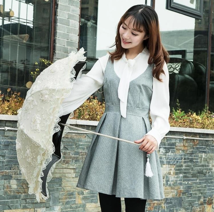 Female model posing with folded lolita parasol in front of stone terrace wall, wearing sweet lolita outfit with grey dress and white blouse, demonstrating portable accessory coordination
