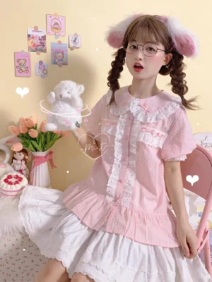 Pink short-sleeve sweet lolita blouse with Peter Pan collar, white lace ruffled trim, and puff sleeves with lace cuffs, worn by model with twin braids in cute pastel-themed seated portrait