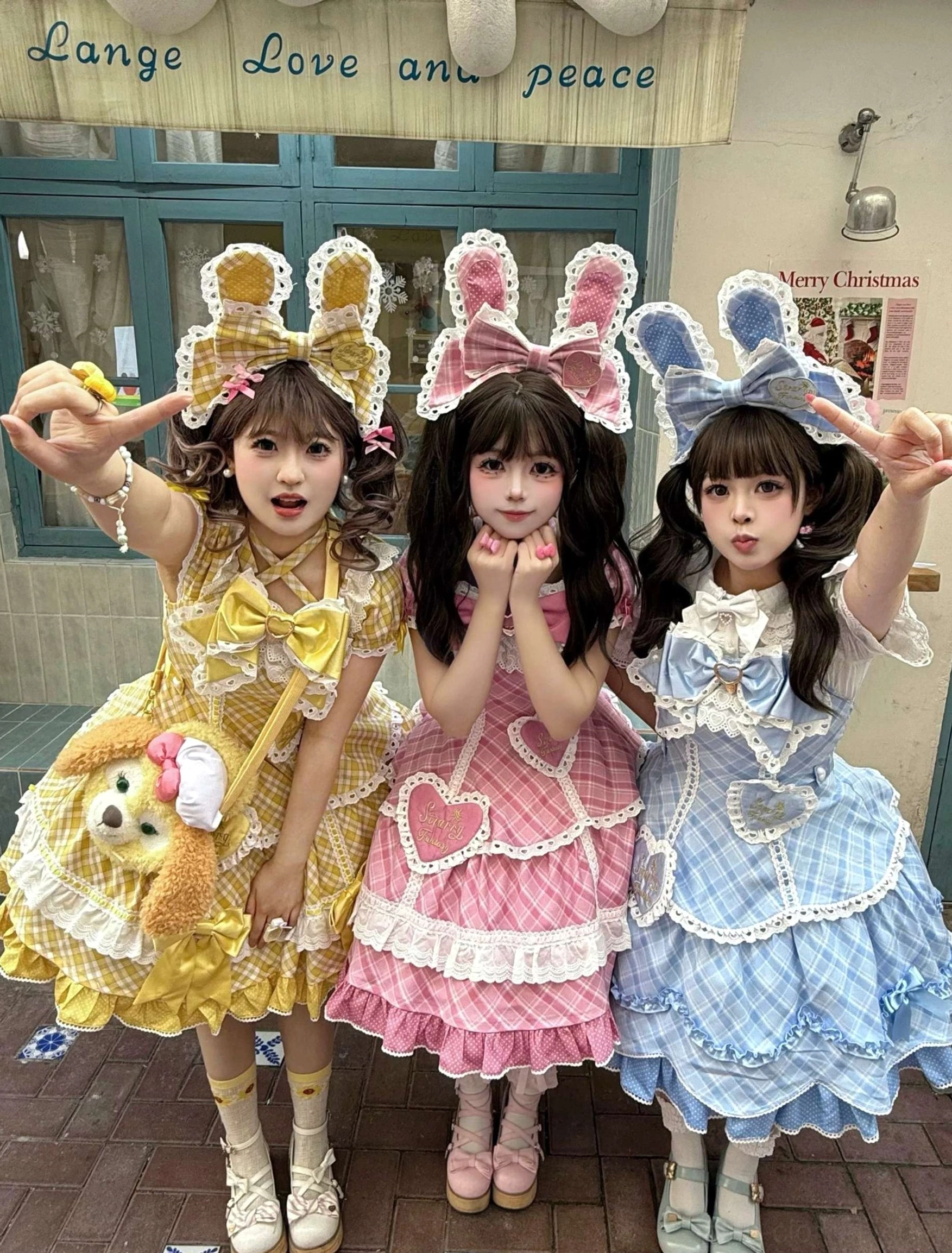 Sweet plaid lolita OP one-piece dresses close-up front full-body triple model view in yellow pink and blue showing oversized bunny ear hair accessories, heart-shaped front patches, tiered ruffled skirt hems with lace accents on retro street storefront backdrop