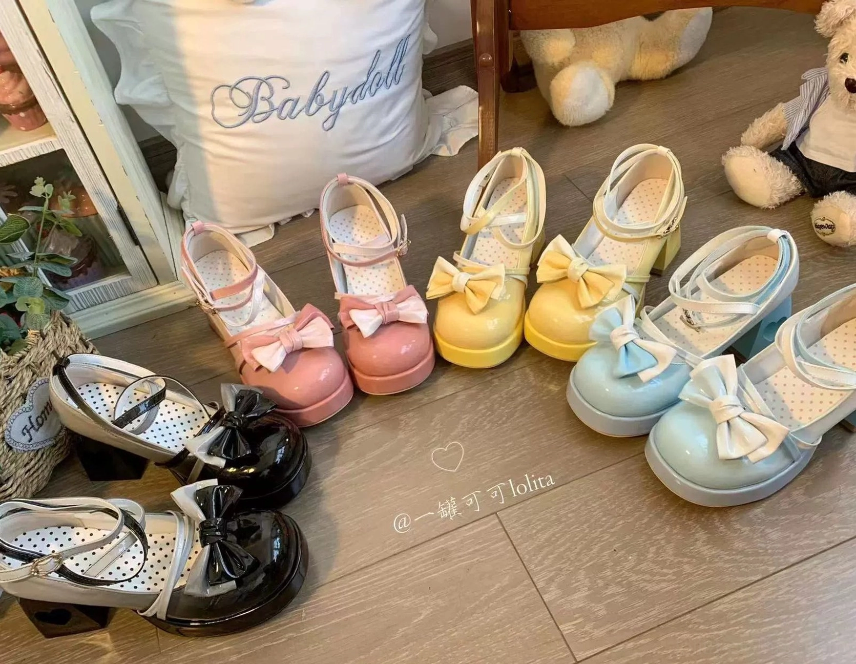 Overhead flat lay collection of assorted Sweet Lolita dress shoes with polished patent leather, bow ankle strap, and heart block heel, arranged with decorative doll and polka dot pillow on wooden floor