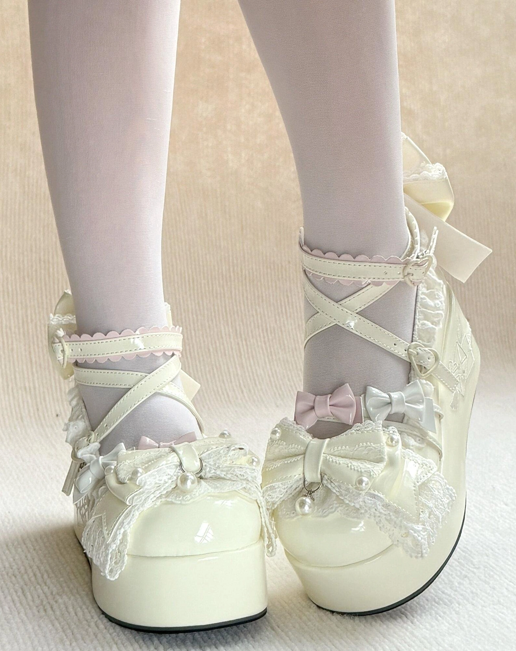 Creamy white off-white Sweet Lolita platform shoes on-foot front view with crisscross straps, ruffled cuff, tiny pink bow embellishment, and glossy off-white patent leather upper styled with white sheer stockings