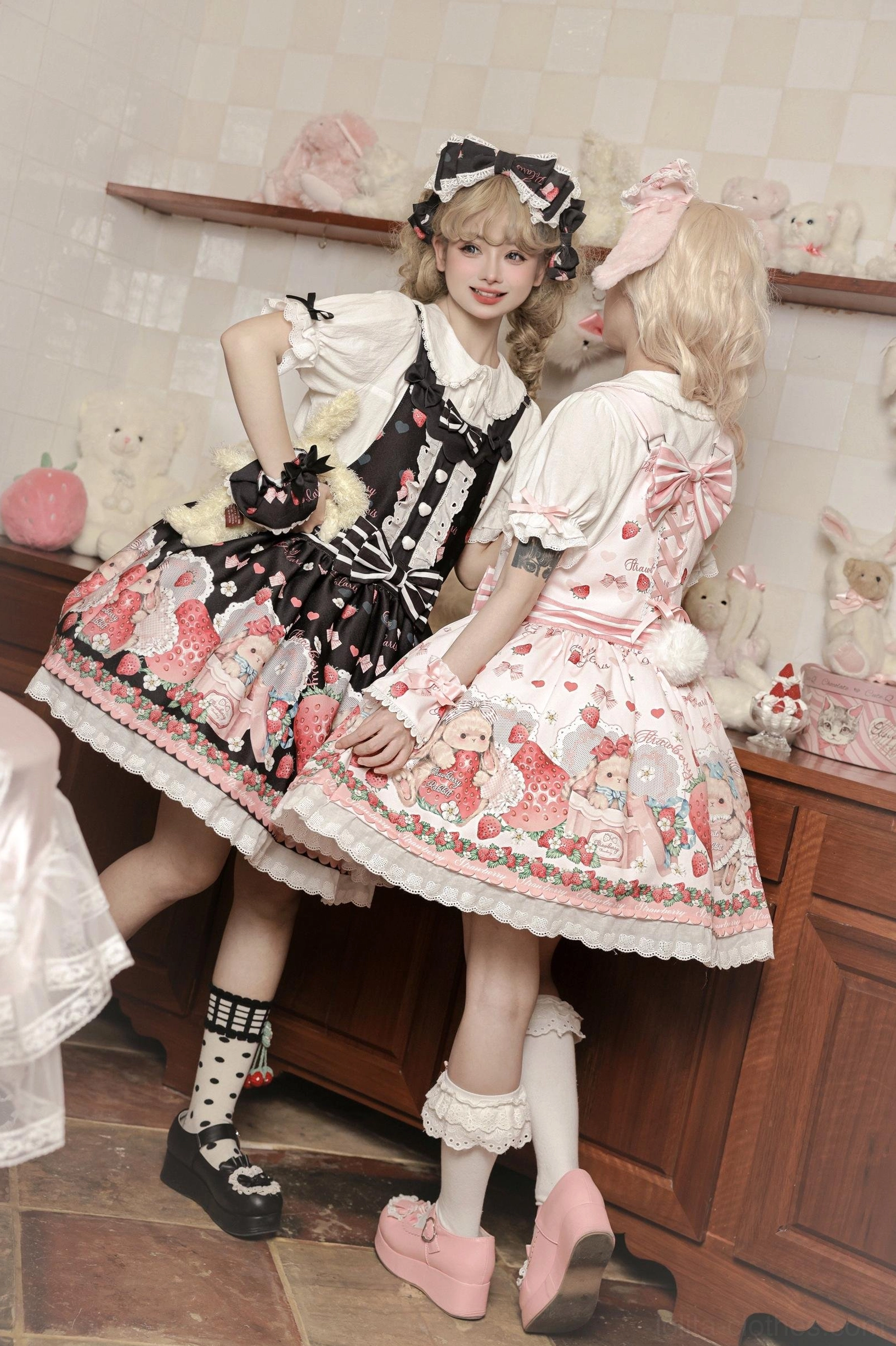 Back side view of two sweet lolita models wearing strawberry print salopette dresses with cross-strap design and decorative bows in vintage kitchen