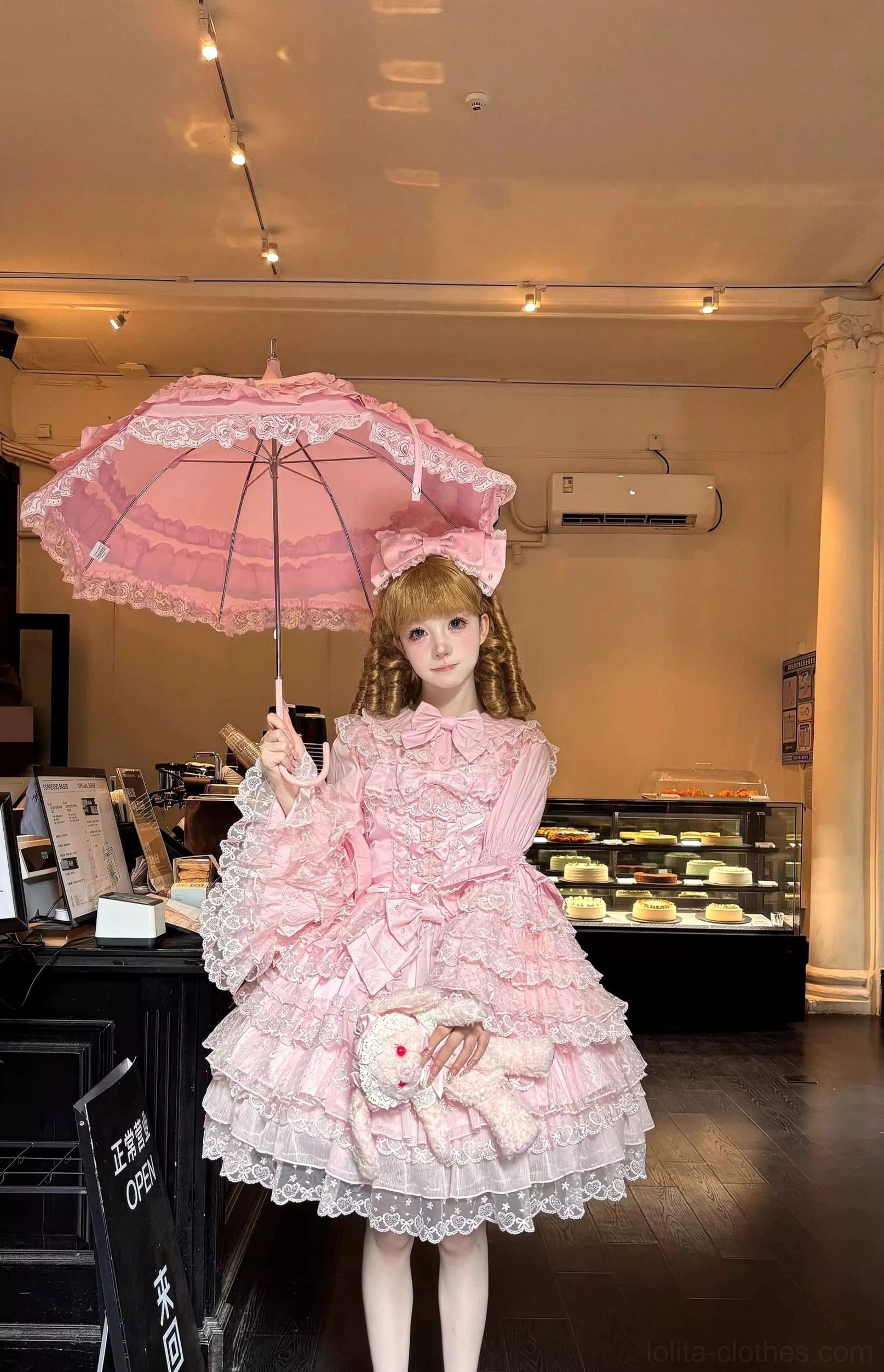 Short length Sweet Lolita dress variation with lace-trimmed skirt, puffed sleeves, and matching pink lace parasol accessory, whimsical bakery setting backdrop