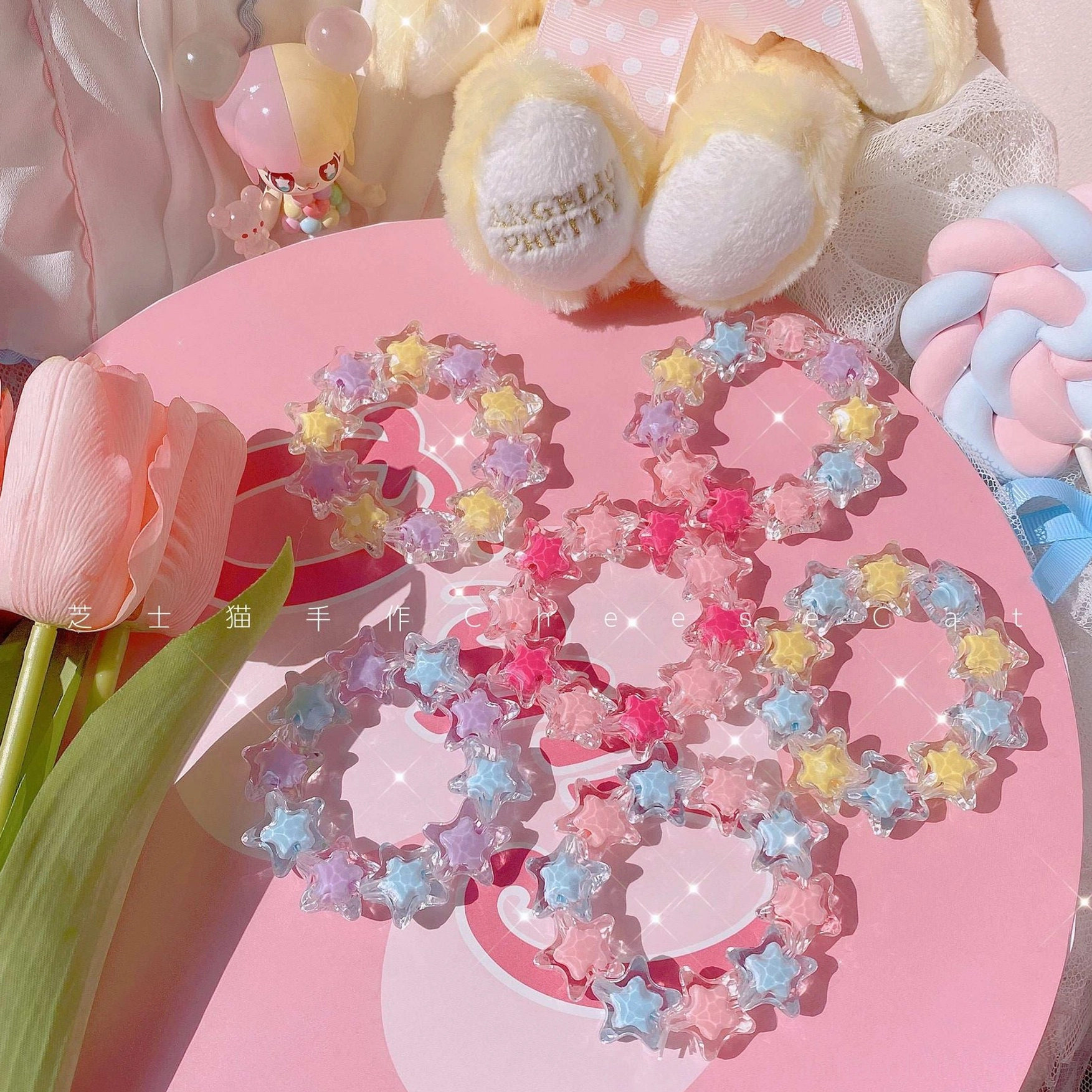 Top-down flat lay display of pastel star-shaped beaded sweet Lolita bracelet set with translucent rainbow acrylic star beads featuring glittery sparkly surfaces, arranged on pink heart-shaped table with plush toys and tulip decorative props