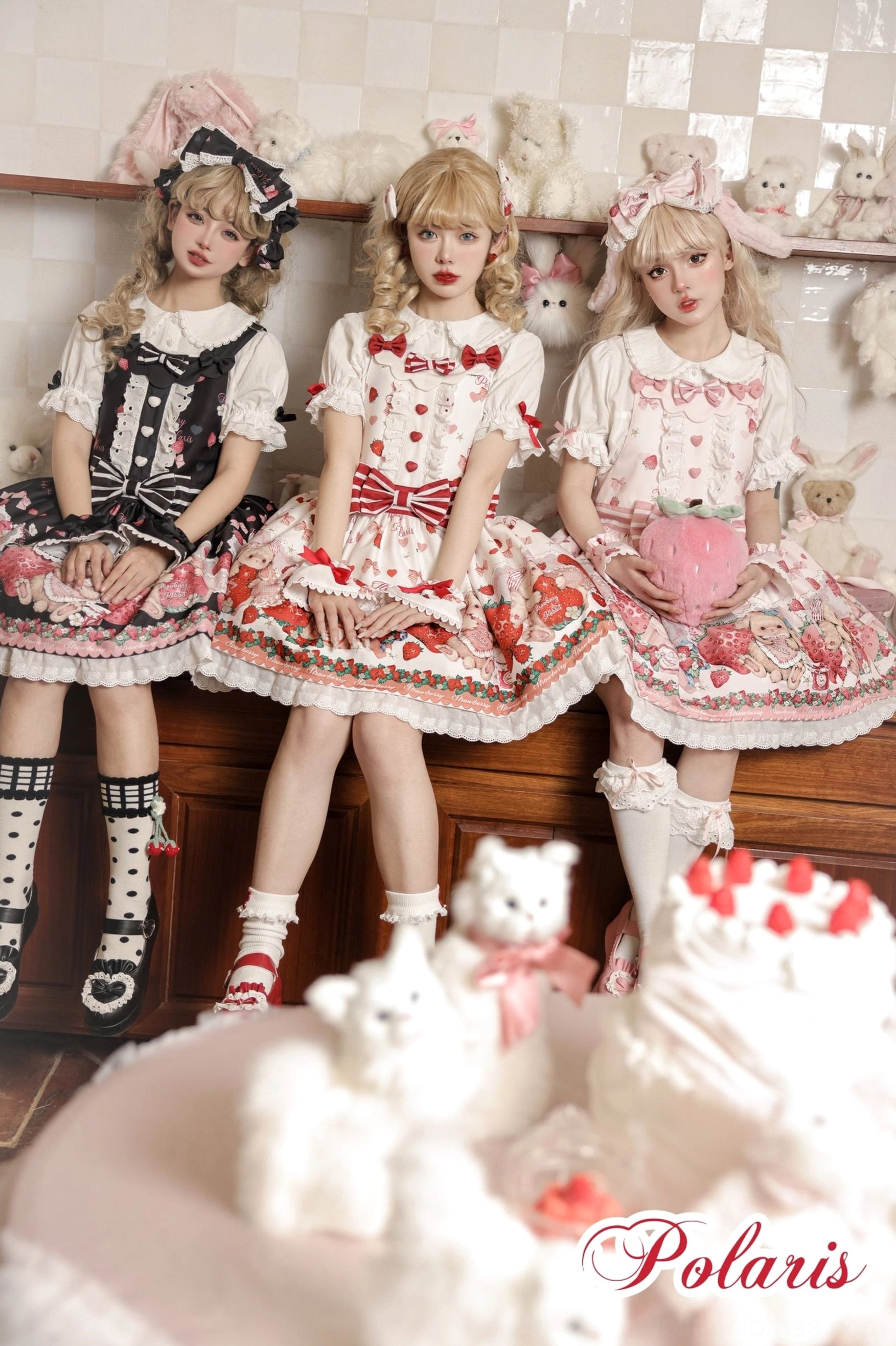 Three blonde sweet lolita models wearing strawberry rabbit print dresses with cake prop in foreground vintage kitchen setting red bow details ruffles